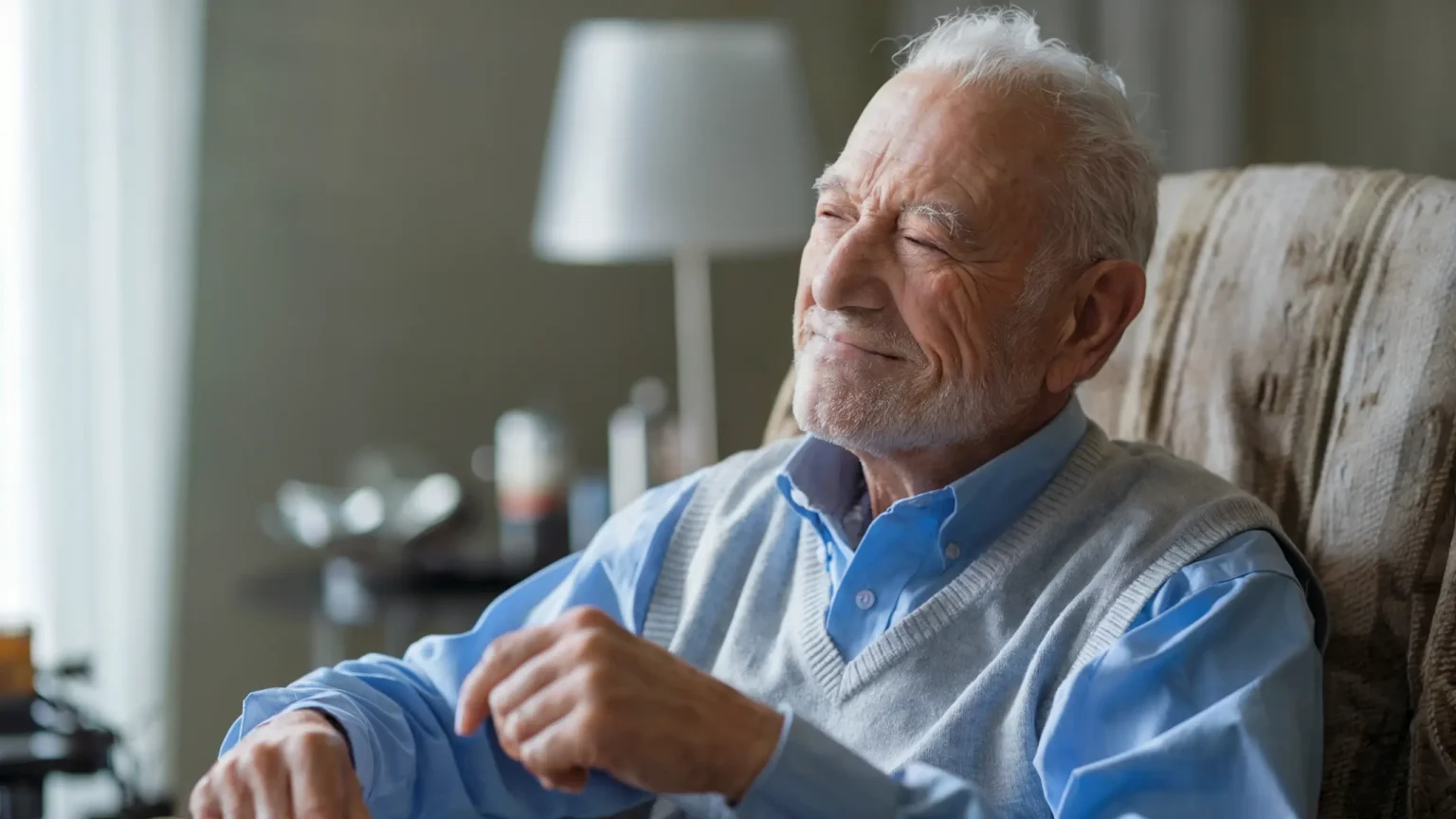 A photo of an elderly man recovering from a stroke. He is sitting in a chair and has a weak smile on his face. He is wearing a blue shirt and a gray vest. He has a white beard and hair. The background is a room with a window, a lamp, and a few items.