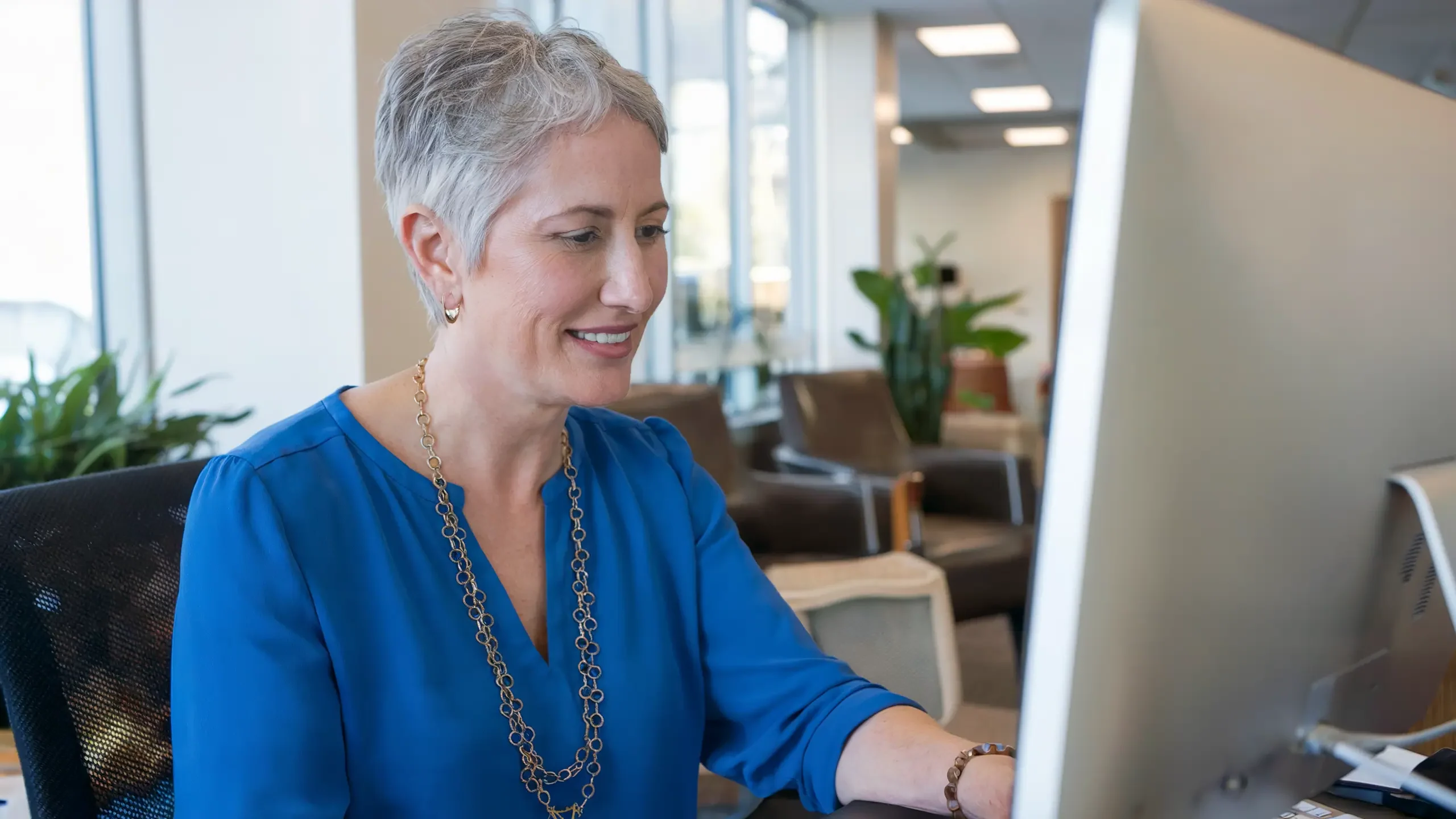 A photo of a senior center director, a woman with short gray hair, wearing a blue blouse and a gold necklace. She is looking at a computer screen. The computer is on a desk in a well-lit room. The room has a few chairs and a plant. The background contains a window.